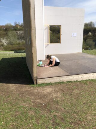 An einem sonnigen Tag sitzt eine Person auf dem Boden neben einer teilweise errichteten Mauer und zeichnet oder schreibt auf Papier. In der Nähe stehen ein Stofftier und eine Wasserflasche.