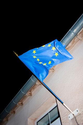Eine Flagge der Europäischen Union mit gelben Sternen auf blauem Grund ist an einem Gebäude angebracht und weht nachts.