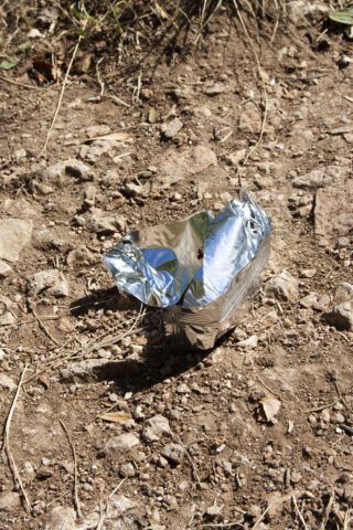 Eine zerknüllte, leere Snacktüte aus Folie liegt weggeworfen auf trockenem, steinigem Boden.