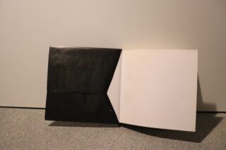 An einer schlichten Wand steht ein geöffneter schwarzer Ordner oder Hefter, auf dessen rechter Seite sich eine leere weiße Seite befindet.