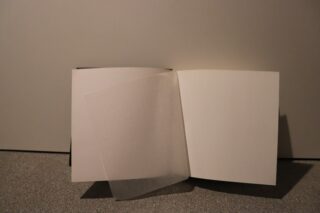 Ein offenes Buch mit leeren Seiten und einer einzelnen, halbtransparenten, teilweise umgeblätterten Seite, die auf einer grauen Oberfläche vor einem schlichten Hintergrund liegt.