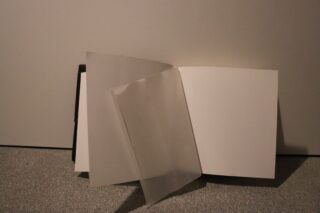 Ein offenes, leeres Buch steht aufrecht an einer Wand. Über die schlichten weißen Seiten ist eine durchsichtige Schutzfolie teilweise geklappt.