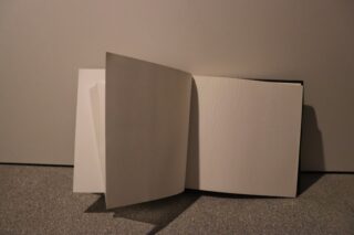 Ein offenes, leeres Buch mit mehreren leeren weißen Seiten steht aufrecht an einer schlichten Wand auf einem Teppichboden.