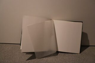 Ein offenes, leeres Buch mit einer teilweise umgeblätterten, durchscheinenden Pergamentseite; das Buch liegt auf einer strukturierten Oberfläche vor einem neutralen Hintergrund.