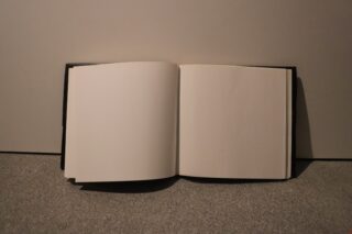 Ein offenes Buch mit leeren Seiten steht aufrecht an einer schlichten Wand auf einem Teppichboden.