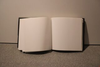 Auf einer flachen, grauen Fläche vor einem schlichten Hintergrund liegt ein offenes, leeres Notizbuch, dessen Seiten völlig leer sind.