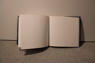 Ein offenes Buch mit leeren, weißen Seiten liegt auf einem grauen Teppich vor einer schlichten Wand.