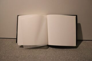 Ein offenes, leeres Buch mit einer durchscheinenden Seite auf der linken Seite, das auf einer flachen, grauen Oberfläche vor einem schlichten Hintergrund liegt.