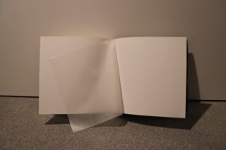 Ein offenes, leeres Buch mit einer durchscheinenden Schutzfolie, die die linke Seite teilweise bedeckt, auf einer grauen Fläche vor einem schlichten Hintergrund.