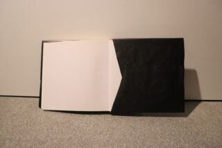 Auf einem grauen Teppich liegt ein aufgeschlagenes Buch, links eine leere weiße Seite, rechts ein schwarzer Einband mit einer Tasche.