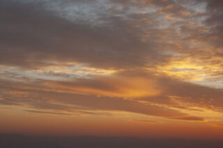Bei Sonnenuntergang ziehen sich orangefarbene und gelbe Wolken über den Himmel, darunter ist der Horizont schwach sichtbar.