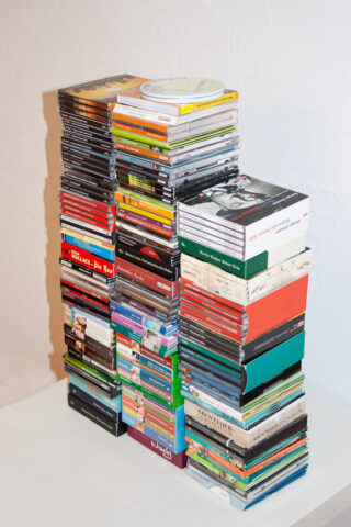 Vier hohe, ungleichmäßige Stapel verschiedenfarbiger CD- und DVD-Hüllen sind auf einer weißen Fläche vor einem schlichten weißen Hintergrund angeordnet.