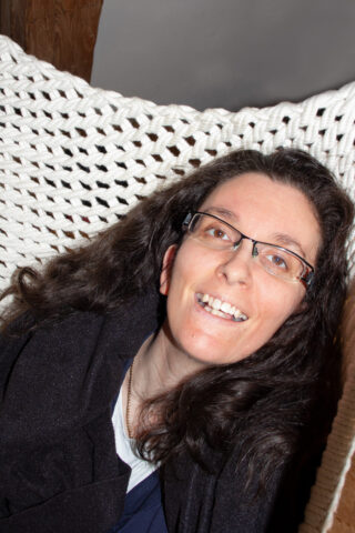 Eine Frau mit langen braunen Haaren und Brille lächelt, während sie auf einer weißen Strickhängematte liegt.