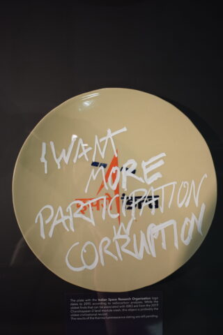 Ein beiges Schild mit dem Logo der Indian Space Research Organisation, darüber der handgeschriebene Text „ICH WILL MEHR PARTIZIPATION UND KORRUPTION.“.