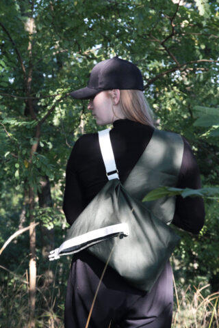 Eine Person mit schwarzer Mütze und schwarzem Outfit steht draußen in einem Waldstück und trägt eine Umhängetasche mit weißem Riemen.