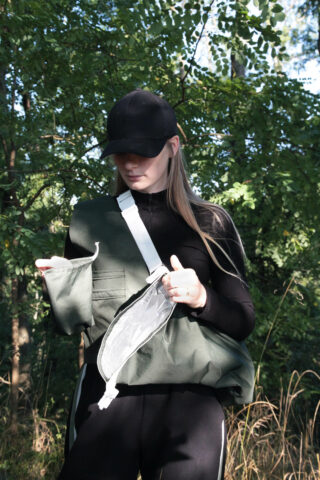 Eine Person mit schwarzer Mütze und Jacke steht im Freien und öffnet eine grüne Tasche mit weißem Riemen. Im Hintergrund sind Bäume und Laub zu sehen.