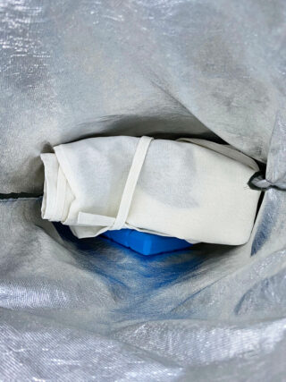 Ein gefaltetes weißes Tuch und ein blauer Plastikgegenstand in einer silberfarbenen Tasche oder einem Behälter.