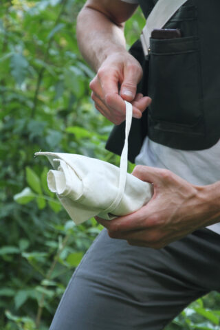 Eine Person im Freien hält ein aufgerolltes weißes Tuch, das mit einem Riemen befestigt ist und von grünem Laub umgeben ist.