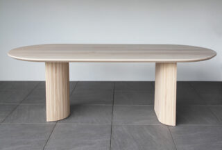 Ein moderner Holztisch mit ovaler Tischplatte und zwei breiten, geschwungenen Beinen, der auf einem gefliesten Boden vor einer schlichten hellen Wand steht.