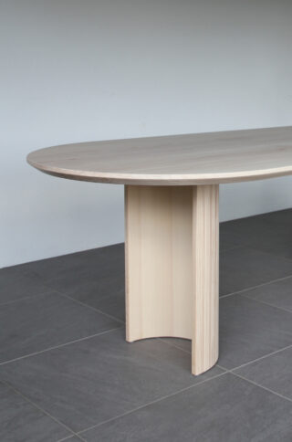 Ein ovaler Tisch aus hellem Holz mit einem einzigen breiten, geschwungenen Mittelbein steht auf einem dunklen Fliesenboden vor einer hellgrauen Wand.