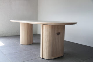 Ein moderner Holztisch mit zwei vertikalen, zylindrischen Beinen und einer glatten, abgerundeten rechteckigen Tischplatte, der auf einem gefliesten Boden vor einer schlichten weißen Wand steht.