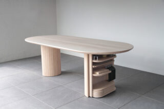 Ein moderner Esstisch aus Holz mit abgerundeten Kanten, zwei dicken Beinen und eingebauten Regalen, auf deren einer Seite sich Küchenutensilien und Tabletts befinden, steht auf einem grau gefliesten Boden.