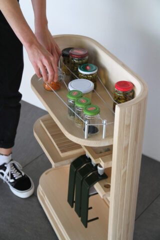 Eine Person ordnet Gewürzgläser auf dem obersten Regal eines Küchenwagens aus hellem Holz an, der über zusätzliche Regale verfügt und mit einer Metallkette gesichert ist.