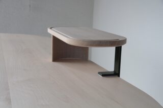 Ein kleines, rechteckiges Holzregal mit gebogener Seite und Metallstütze ist auf einer hellen Holzoberfläche an einer schlichten Wand montiert.