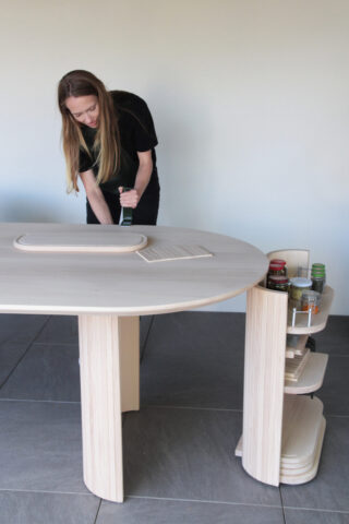 Eine Person verwendet ein Küchengerät an einem hellen Holztisch mit eingebauten Regalen, auf denen Gläser und Behälter auf gefliestem Boden stehen.