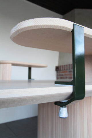 Nahaufnahme eines hellen Holztisches mit einer an der Kante befestigten grünen Metallklammer; im Hintergrund ist ein weiterer ähnlicher Tisch zu sehen.