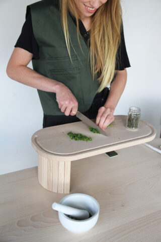 Eine Person mit langem blondem Haar hackt Kräuter auf einem erhöhten Holzschneidebrett. Auf dem Tisch daneben stehen ein Glas mit Kräutern sowie ein Mörser mit Stößel.