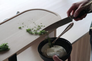 Eine Person schaben gehackte Kräuter mit einem Messer von einem Schneidebrett in eine schwarze Schüssel mit weißem Joghurt oder Sauerrahm.