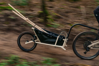 Ein Fahrrad mit einem angehängten einrädrigen Lastenanhänger bewegt sich auf einem Feldweg in einem Waldgebiet.