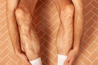 Eine Person mit haarigen Beinen und weißen Socken sitzt auf einem Fischgrätfliesenboden und hält ihre Beine. An einem Schienbein sind blaue Perlen befestigt.