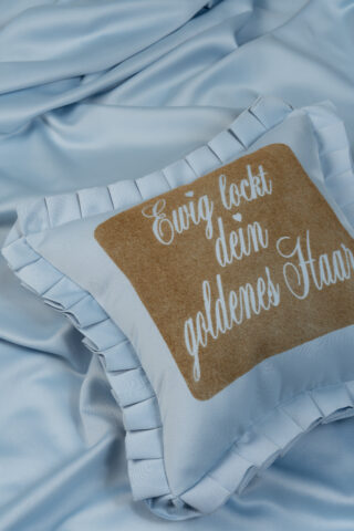 Ein hellblaues Kissen mit gerüschten Rändern und einem hellbraunen Quadrat mit den deutschen Worten „Ewig lockt dein goldenes Haar“, das auf einem passenden blauen Stoff ruht.