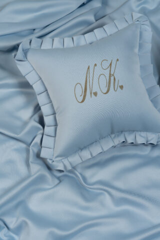 Hellblaues Kissen mit gerüschten Rändern und den goldgestickten Initialen „N.K.“ liegt auf passendem Satinstoff.