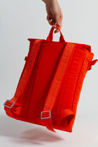 Eine Hand hält einen orangefarbenen Rucksack am oberen Griff und zeigt die gepolsterten Schultergurte und Schnallen vor einem schlichten weißen Hintergrund.