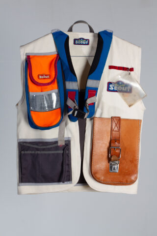 Eine beige Pfadfinderweste mit mehreren Taschen in Blau, Orange, Braun und Grau und einem Namensschild im Brustbereich.