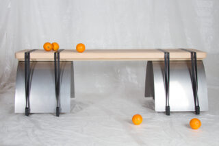 Auf einer weißen Fläche steht eine moderne Bank mit Metall- und Holzelementen, auf und um die Bank herum sind mehrere Orangen platziert.