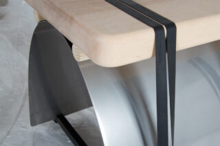 Eine Nahaufnahme einer modernen Tischecke mit einer hellen Holzplatte, einem schwarzen Metallrahmen und einer gebogenen silbernen Metallstütze darunter.