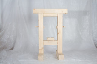 Eine Holzkonstruktion mit zwei vertikalen Balken und einem horizontalen Balken oben, vor einem Hintergrund aus durchscheinenden Kunststoffplatten.