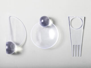 Auf einer hellen Fläche sind drei durchscheinende Plastikutensilien mit runden, gräulichen Griffen nebeneinander angeordnet; eines ist eine Gabel und zwei sind löffelartig.