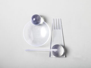 Ein Teller, eine Gabel und ein Messer aus durchsichtigem Kunststoff sind mit zwei durchscheinenden Kugeln auf einer hellgrauen Oberfläche angeordnet.