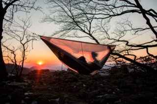 Eine Person liegt in einer Hängematte, die mit einem Maschennetz bedeckt ist, zwischen Bäumen aufgehängt ist und den Sonnenuntergang in einer felsigen Umgebung im Freien beobachtet.