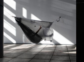 Eine Person liegt in einer halbtransparenten Hängematte im Innenbereich, wobei das Sonnenlicht geometrische Schatten auf den gefliesten Boden und die weiße Wand wirft.