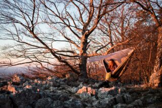 Eine Person sitzt bei Sonnenuntergang in einer Hängematte, die zwischen Bäumen auf einem Felsvorsprung aufgehängt ist, umgeben von blattlosen Zweigen und beleuchtet vom warmen Sonnenlicht.