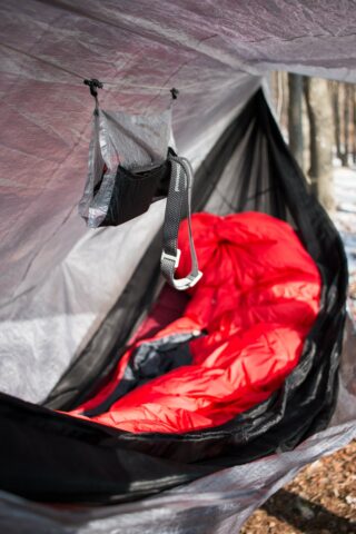 Ein roter Schlafsack liegt in einer schwarzen Hängematte unter einer grauen Plane. Darüber hängt ein kleiner Beutel mit Riemen. Im Hintergrund sind Bäume und Schneeflecken zu sehen.