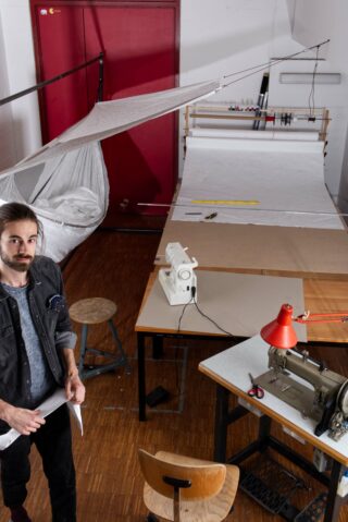 Ein Mann steht in einer Werkstatt mit Nähmaschinen, großen Stoffbahnen, einer roten Tür und verschiedenen Werkzeugen auf Arbeitstischen.