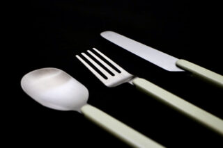 Auf schwarzem Untergrund sind Löffel, Gabel und Messer mit hellgrünen Griffen diagonal angeordnet.