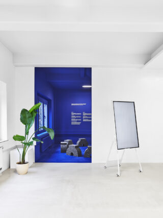 Ein minimalistischer weißer Raum mit einer Topfpflanze, einer leeren Staffelei und einer Tür, die den Blick auf einen Bereich mit blauen Wänden, Steinsitzen und Wandtext freigibt.